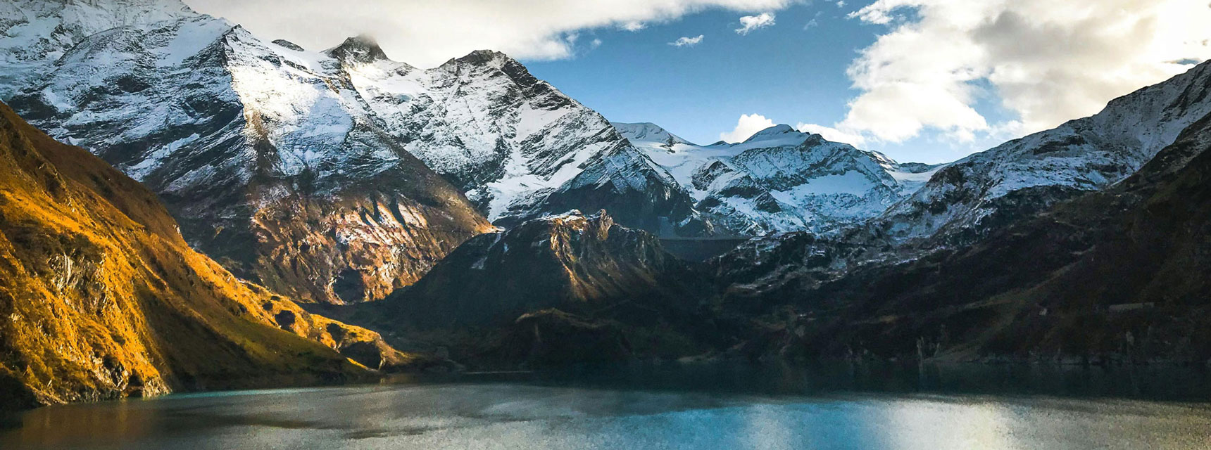 Austrian snow-capped mountains and hillsides reflected in a calm lake, under a partly cloudy sky.