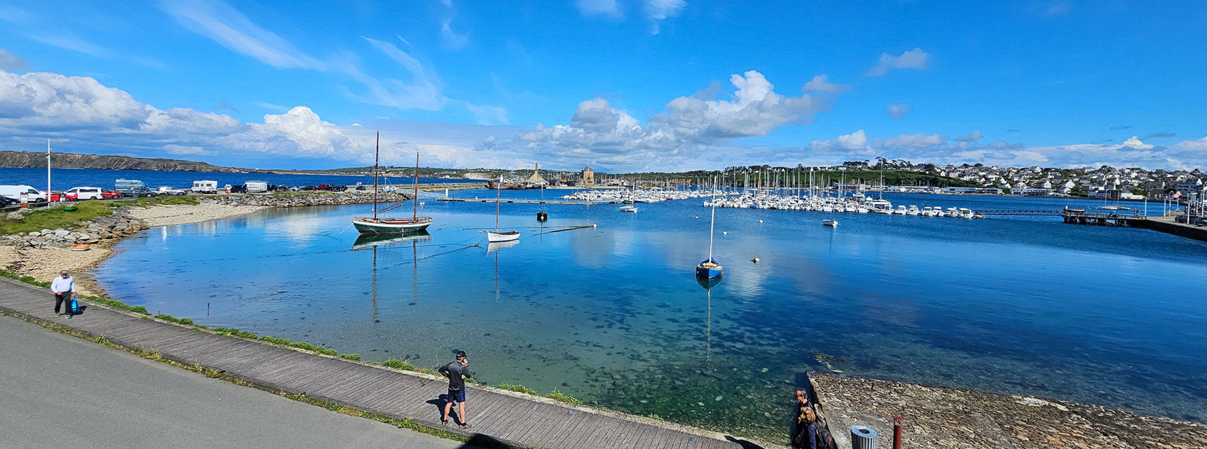 A calm harbor in Brittany with sailboats on clear blue water, under a bright sky with scattered clouds.